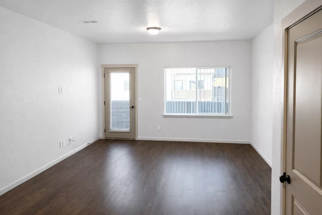 Empty room with white walls, brown wooden floor, a window with blinds, and a door with a glass panel.