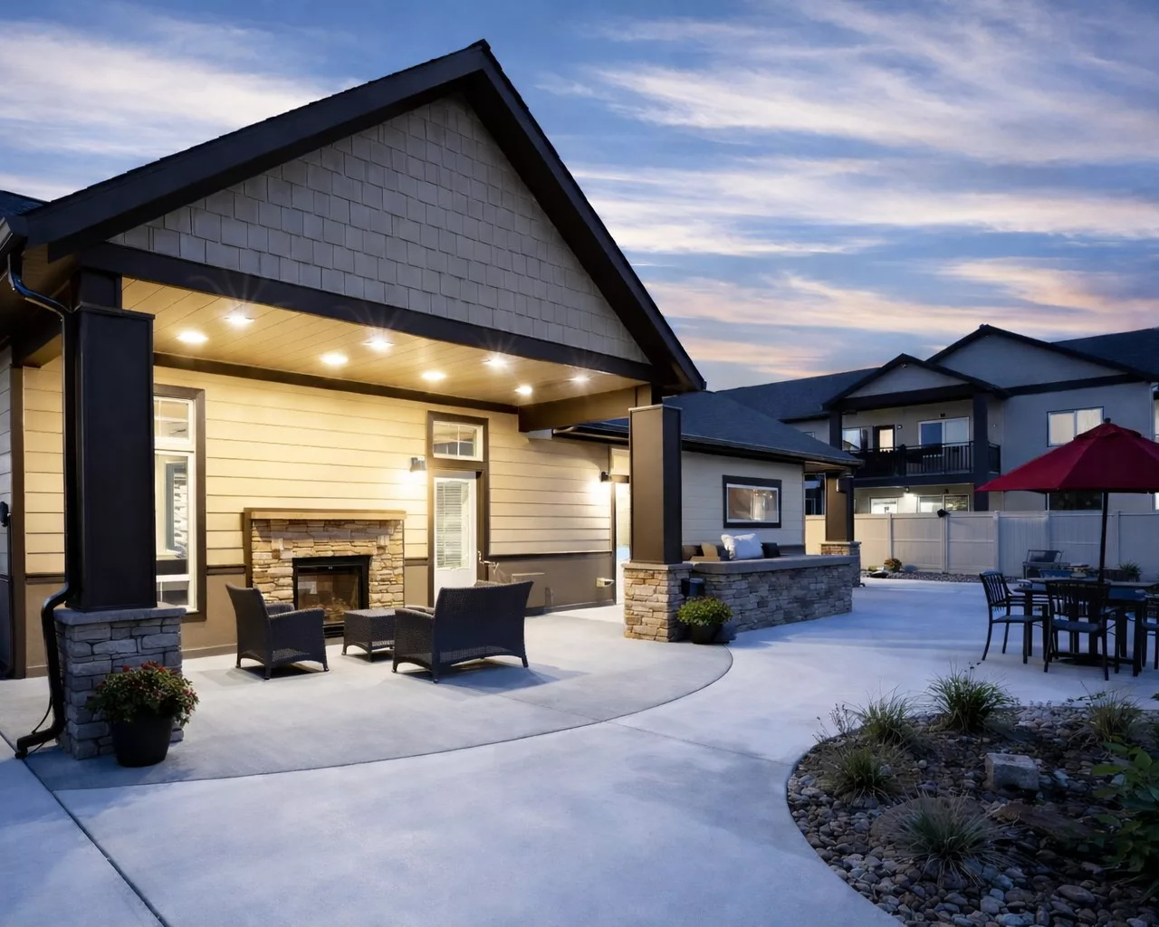 Modern patio at dusk with cozy seating, a stone fireplace, and an outdoor dining area under a red umbrella.