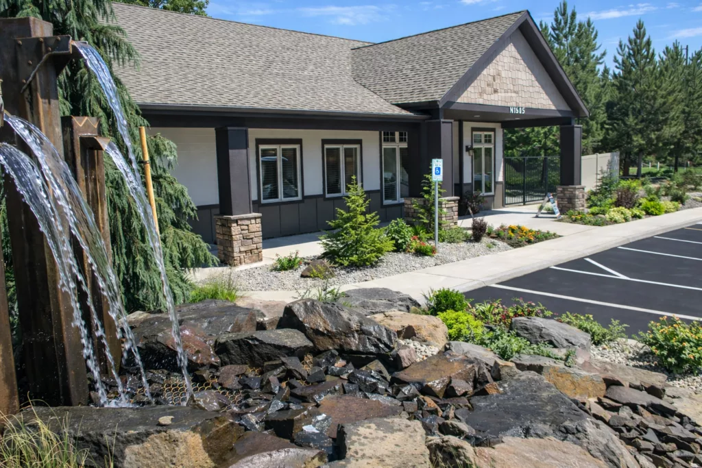 A building with stone accents features a rock waterfall in the foreground and neatly landscaped surroundings.