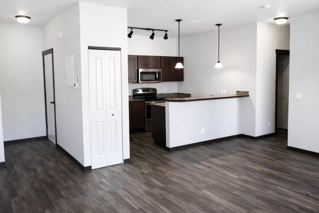 Modern kitchen and living area with wood flooring, white walls, dark cabinets, and stainless steel appliances under pendant lighting.