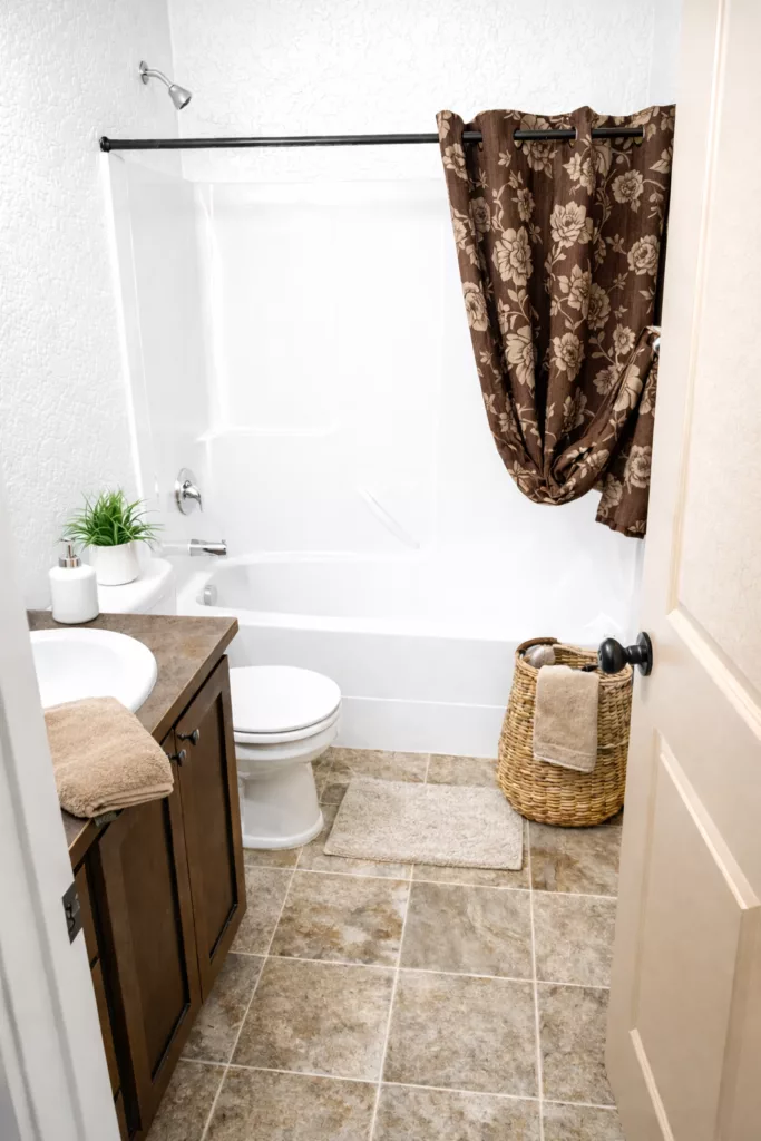 A small bathroom with a bathtub, brown floral shower curtain, white toilet, sink with wooden cabinetry, and wicker basket.
