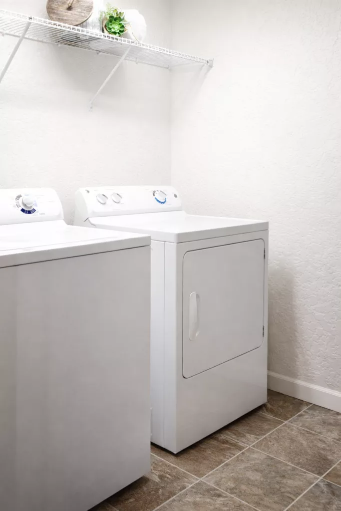 A laundry room with a white washing machine and dryer on a tiled floor, under an empty wire shelf.