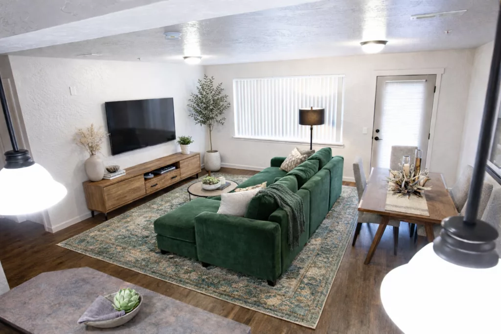 A modern living room with a green sectional sofa, a flat-screen TV on a wooden stand, and a dining table with decorative items.