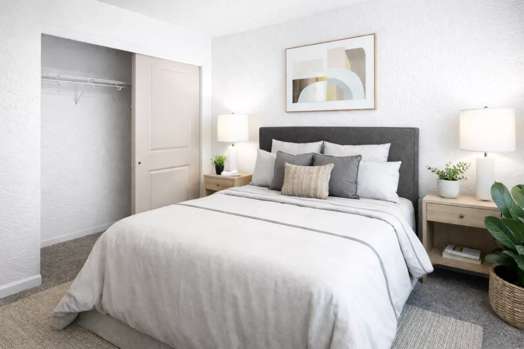 A neatly arranged bedroom with a double bed, gray upholstered headboard, matching nightstands with lamps, and a sliding closet door.