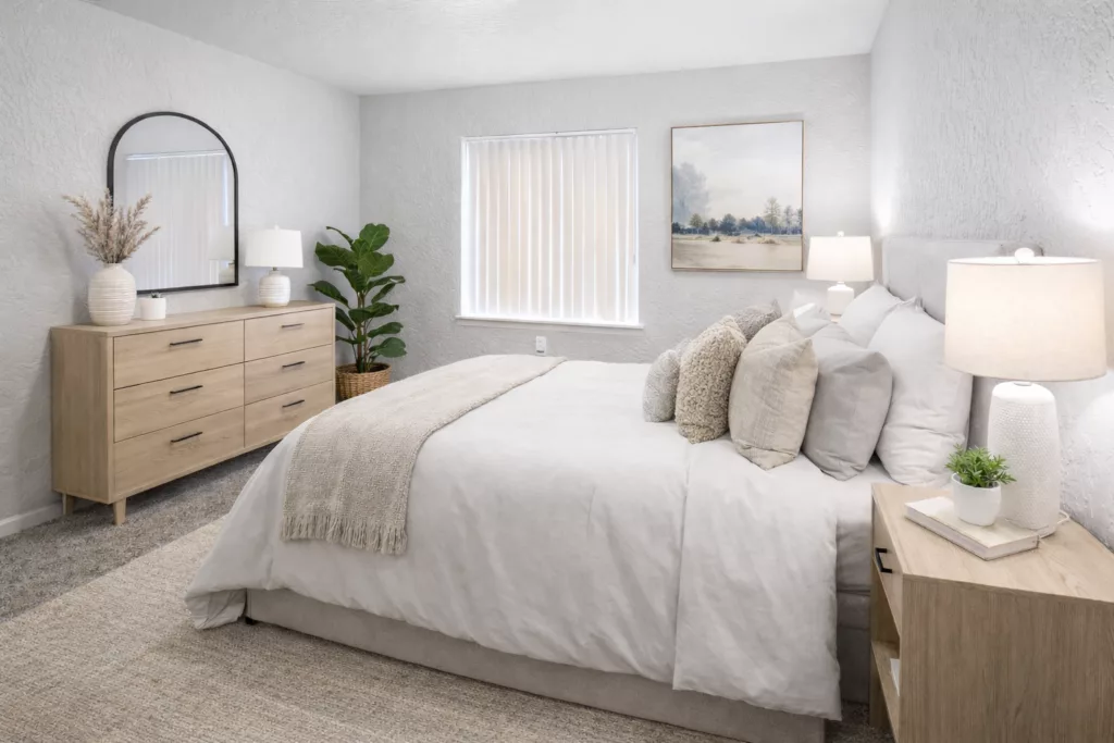A cozy bedroom with a white bed, light wood furniture, a round mirror, and plant decor, softly lit by table lamps.