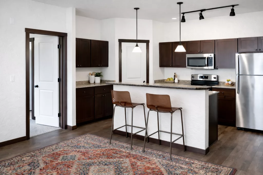 A modern kitchen with dark wooden cabinets, stainless steel appliances, a bar counter with two brown stools, and a patterned rug.