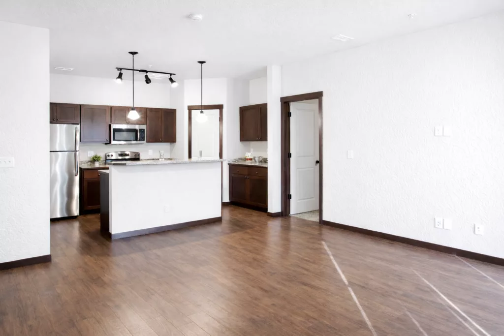 Empty modern kitchen and living area with wooden floors, white walls, dark cabinetry, stainless steel appliances, and pendant lighting.