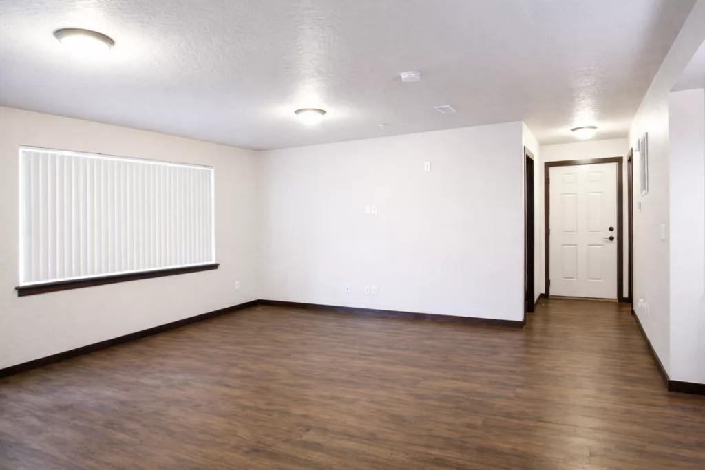 An empty room with white walls, a wooden floor, ceiling lights, a window with blinds, and a white door.