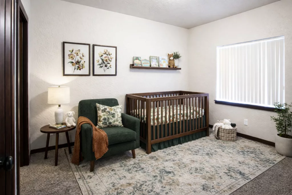 A cozy nursery with a wooden crib, green armchair, shelf with books, and floral decor on the walls.
