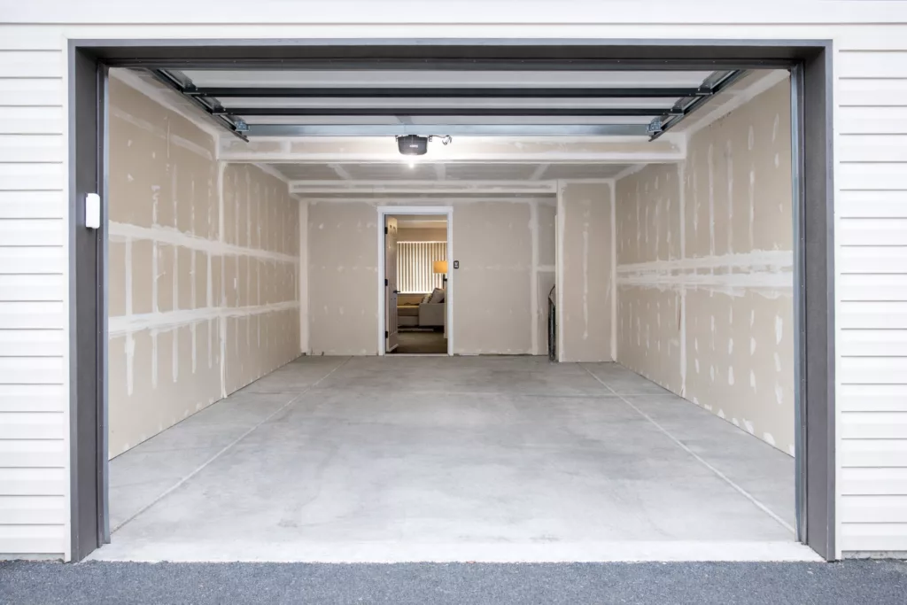 Empty, freshly constructed garage with unfinished drywall and an open entrance leading to an interior room.