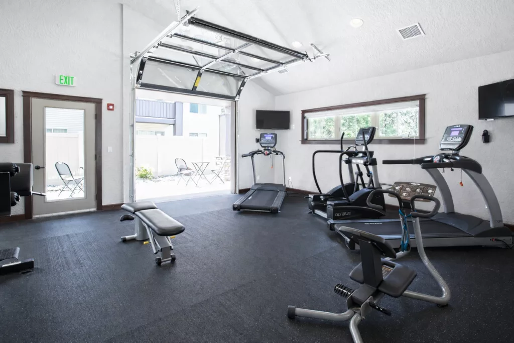 A gym with exercise equipment including treadmills, an exercise bike, and benches, with a door open to an outdoor area.